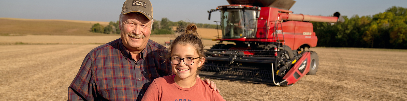 A man and his granddaughter in a harvested field with combine in the background. 